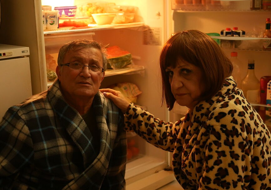 Two people in bathrobes sit in front of an open refrigerator. 