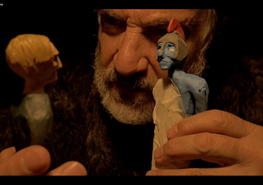 up close shot of a man holding two clay-like figurines. One figurine is blue with a white tunic. 