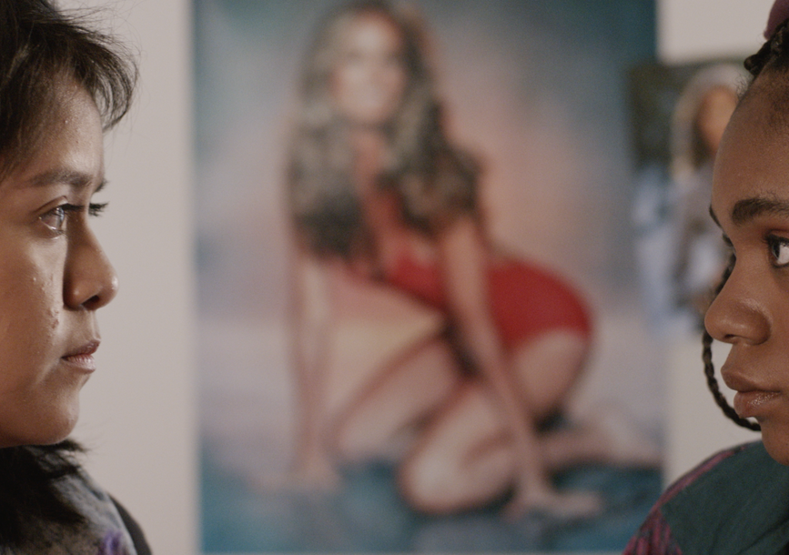 Film still from Gently Jennifer depicting two girls looking at one another with a poster of a woman between them.