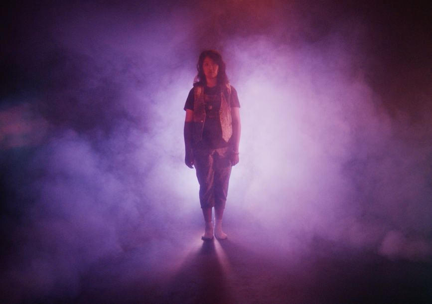 Film still from Gently Jennifer depicting a girl in spotlight surrounded by smoke.