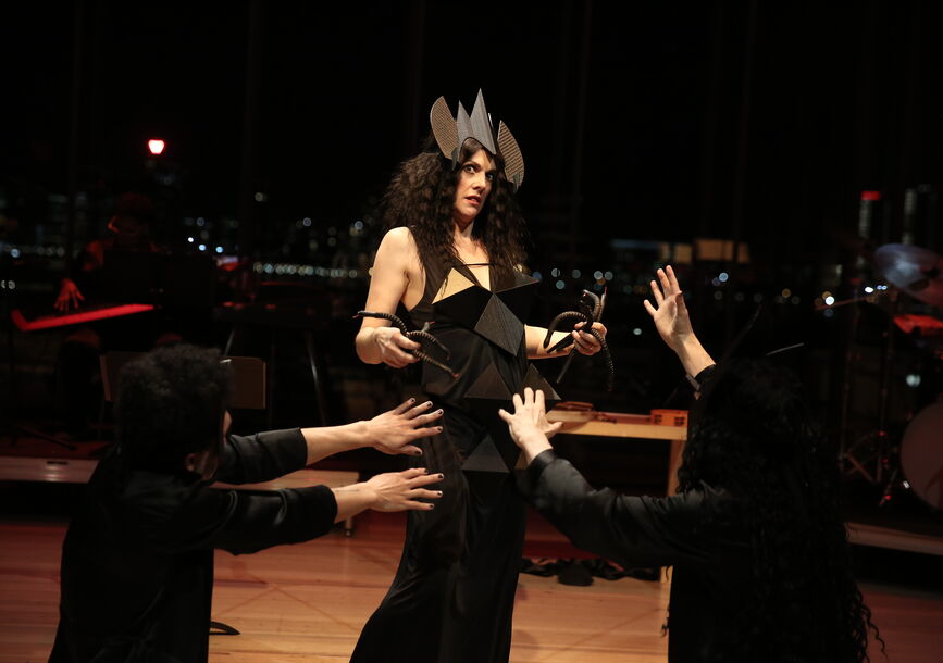 A woman stands in a black dress while two men kneel before her.