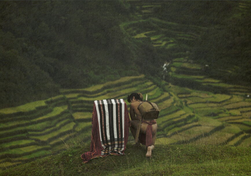 A person crouches on top of a green hill.
