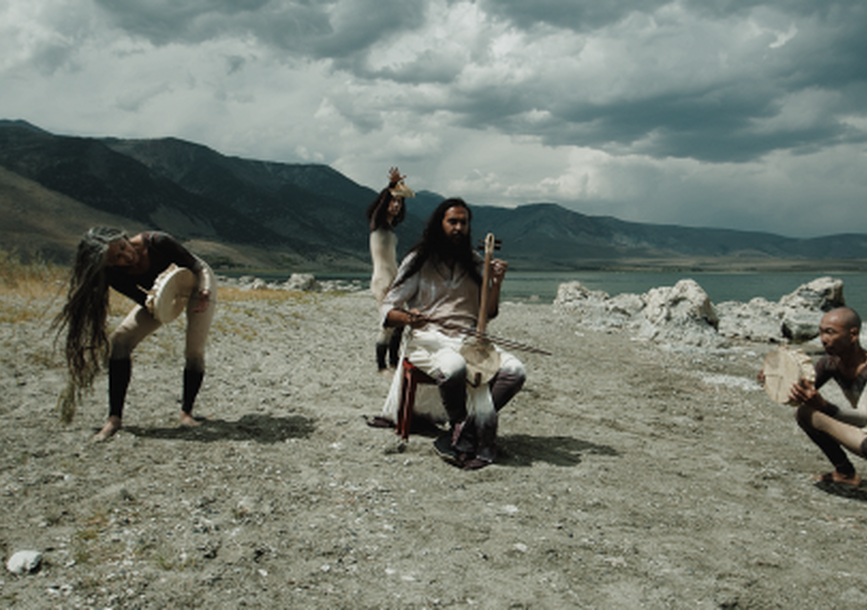 4 figures on a dirt ground, performing a tribal dance and chant