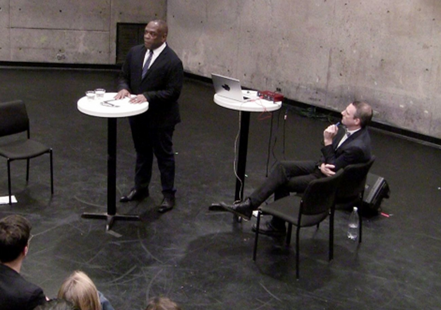 2 men on stage - one standing giving a speech and the other sitting casually in front of people 