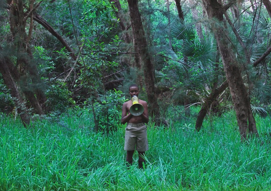 A person stands with a megaphone in the middle of a forest.