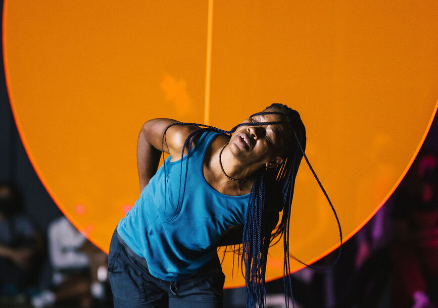 Okwui dances in front of a large orange circle.