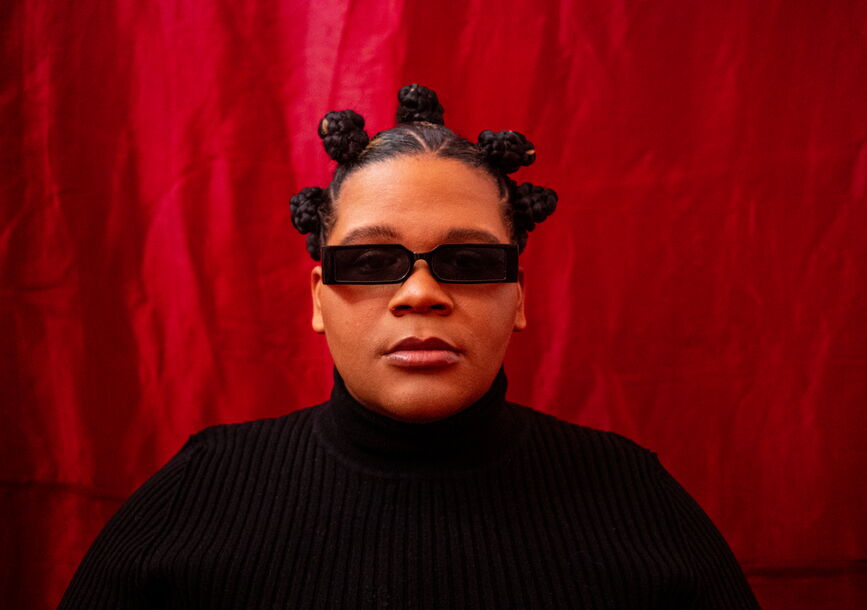 Joy wears sunglasses in front of a red background.