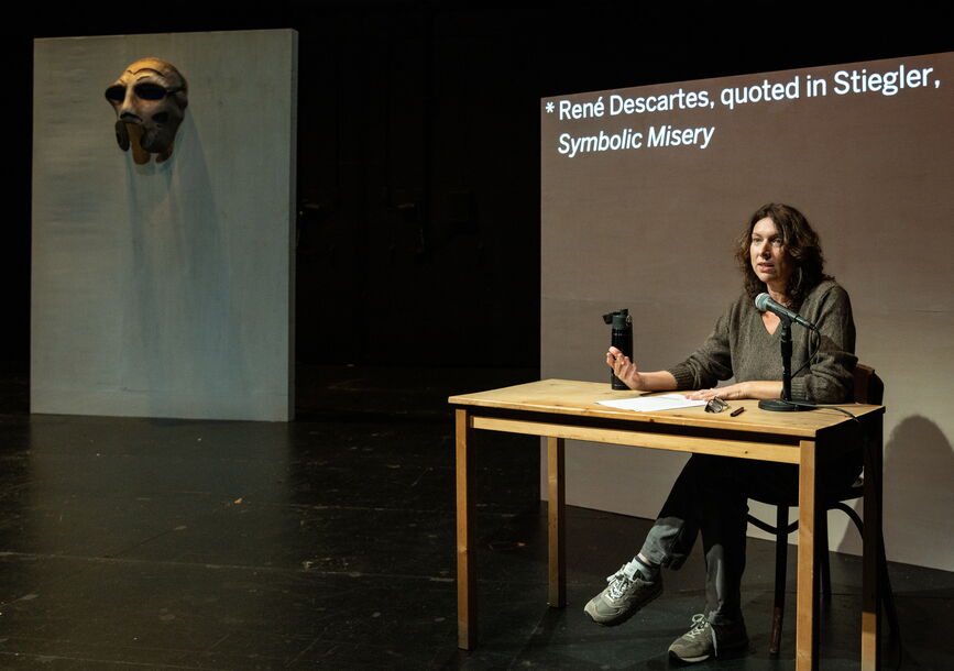 Annie Dorsen sits at a table in front of a projection screen.
