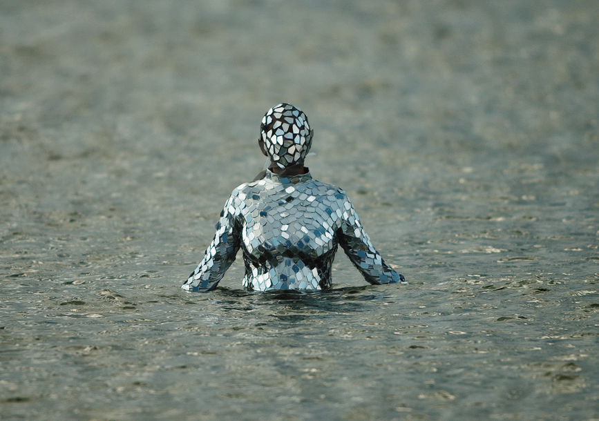 humanoid figure dressed in a outfit with slats of mirror pieces covering all over; coming out of a body of water