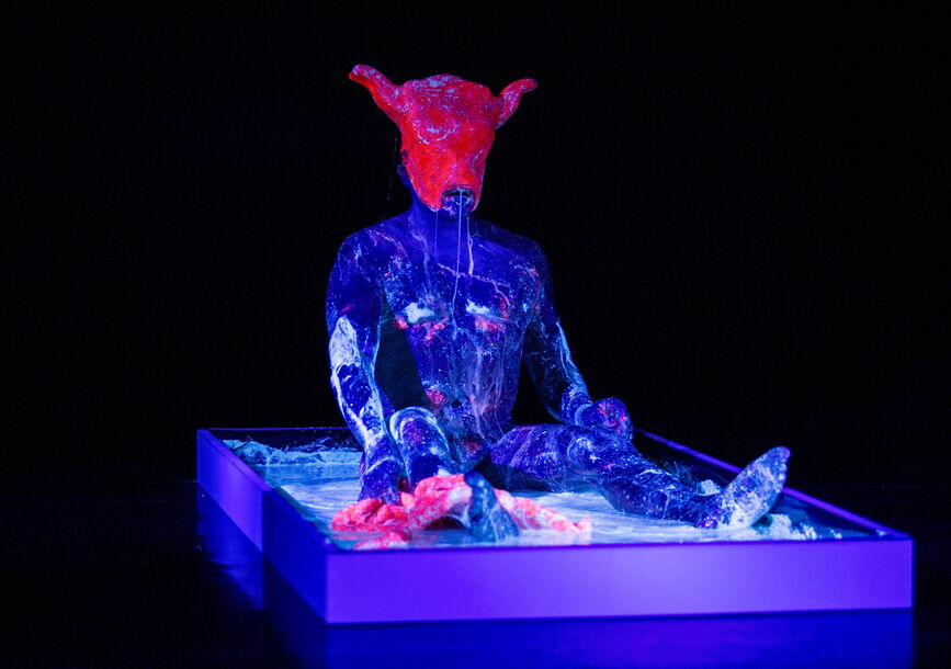 humanoid figure sitting up on a platform, covered in purple paint visible in a black light. figure is wearing a pink like hat with cow ears or horns