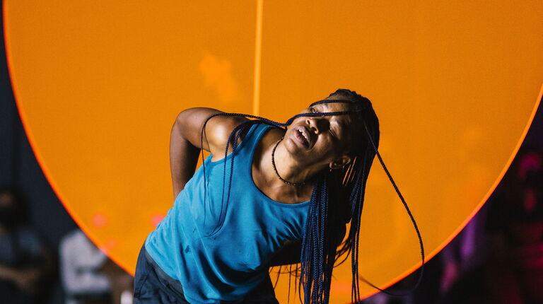 Okwui dances in front of a large orange circle.