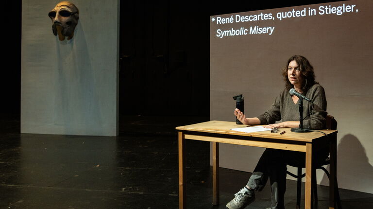 Annie Dorsen sits at a table in front of a projection screen.