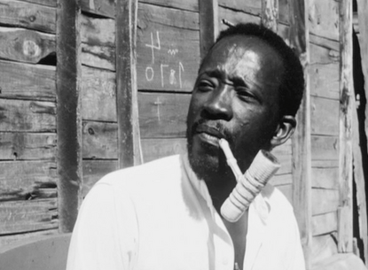 A person in front of a wooden wall with a pipe in their mouth.