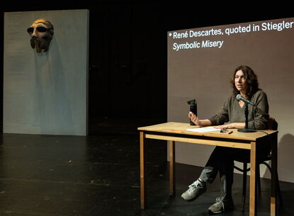 Annie Dorsen sits at a table in front of a projection screen.