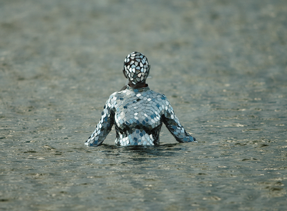 humanoid figure dressed in a outfit with slats of mirror pieces covering all over; coming out of a body of water