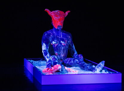 humanoid figure sitting up on a platform, covered in purple paint visible in a black light. figure is wearing a pink like hat with cow ears or horns