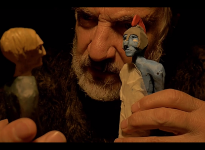 up close shot of a man holding two clay-like figurines. One figurine is blue with a white tunic. 